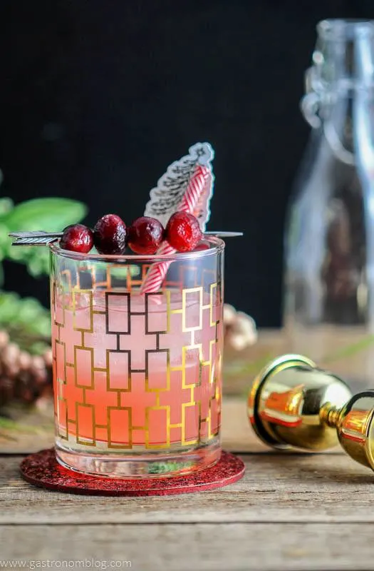 The Red Hound Cocktail in a rocks glass and garnished with cranberries and straws, on a wooden table.