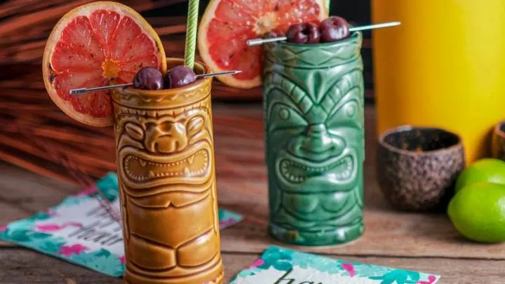 a pair of tiki glasses filled with The Rambler's Ruby, a tiki cocktail garnished with brandied cherries and a bruleed grapefruit.