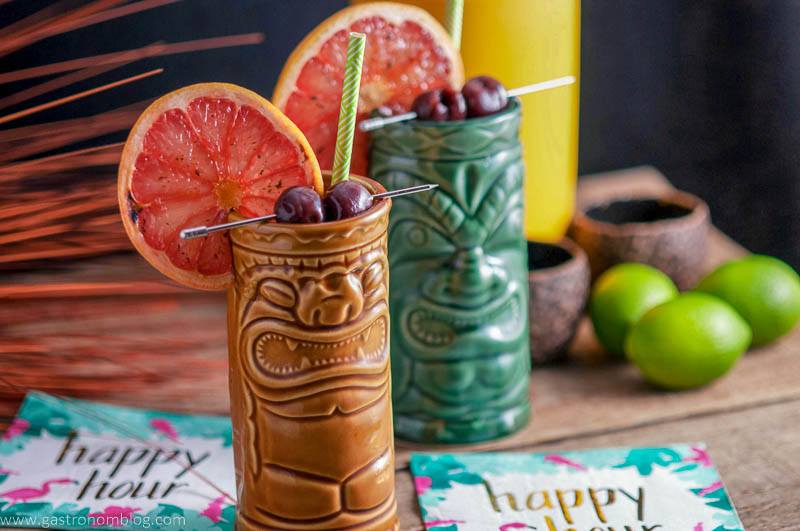 A pair of tiki glasses filled with The Rambler's Ruby, a tiki cocktail garnished with brandied cherries and a bruleed grapefruit.