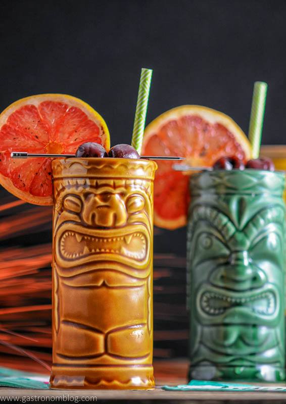 Close up of a pair of tiki glasses filled with The Rambler's Ruby, a tiki cocktail garnished with brandied cherries and a bruleed grapefruit.