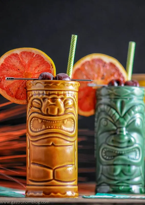 Close up of a pair of tiki glasses filled with The Rambler's Ruby, a tiki cocktail garnished with brandied cherries and a bruleed grapefruit.