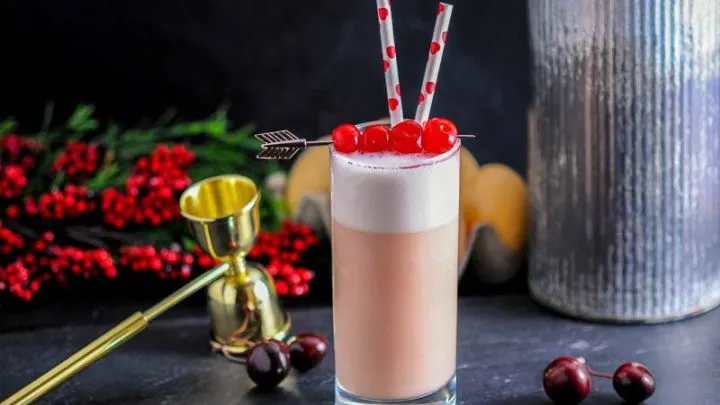 A collins glass sits garnished by maraschino cherries a top a rich foam filled Cherry Toasted Cream Ramos Gin Fizz