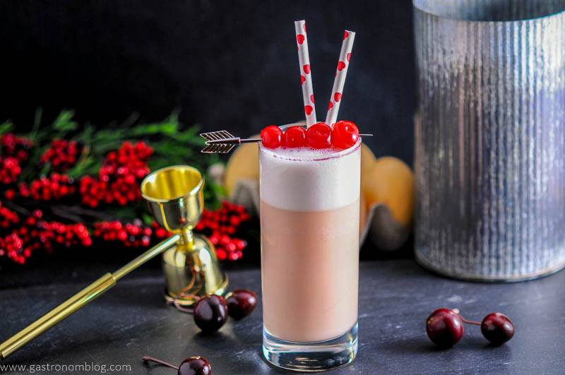 A collins glass sits garnished by maraschino cherries a top a rich foam filled Cherry Toasted Cream Ramos Gin Fizz