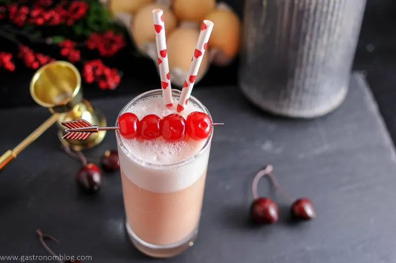 A highball glass containing the cherry toasted cream ramos gin fizz cocktail with maraschino cherries for a garnish and doecrative straws.