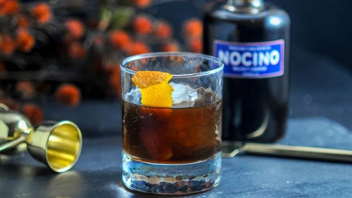 The Nutty Rum Old Fashioned cocktail in a rocks glass with ice ball and orange peel garnish and bottle of Nocino Walnut Liqueur from Prohibition Spirits.