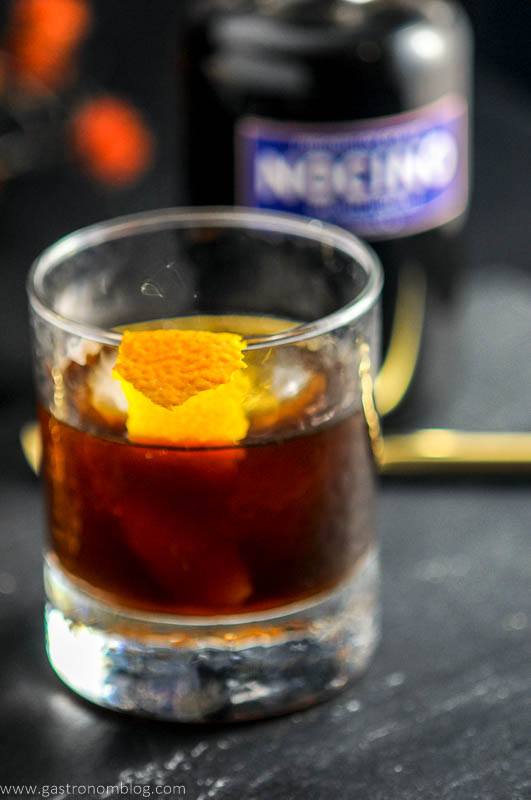 THe Nutty Rum Old Fashioned cocktail in a rocks glass with ice ball and orange peel garnish and bottle of Nocino Walnut Liqueur from Prohibition Spirits.