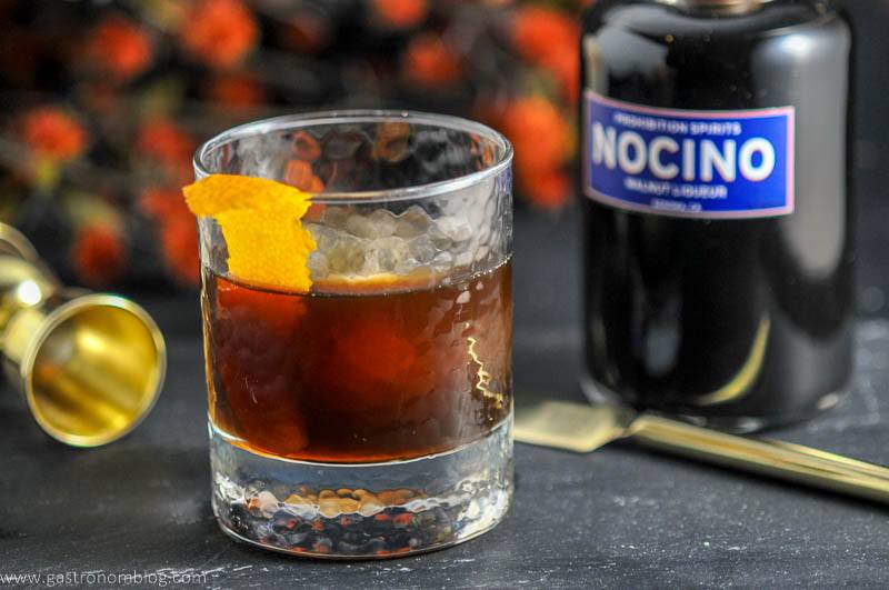 THe Nutty Rum Old Fashioned cocktail in a rocks glass with ice ball and orange peel garnish and bottle of Nocino Walnut Liqueur from Prohibition Spirits.