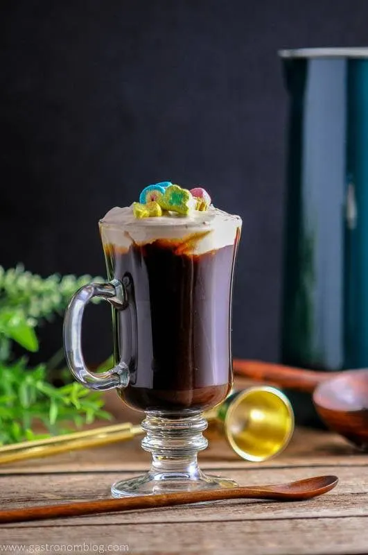 Coffee in glass mug with cream top and Lucky Charms Cereal on top. On a wooden table with wooden spoon, greenery and gold barware behind