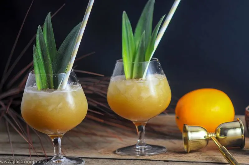 Coconut Rum Painkillers in brandy glasses with pineapple fronds and straws. Orange, ounce measure in background.