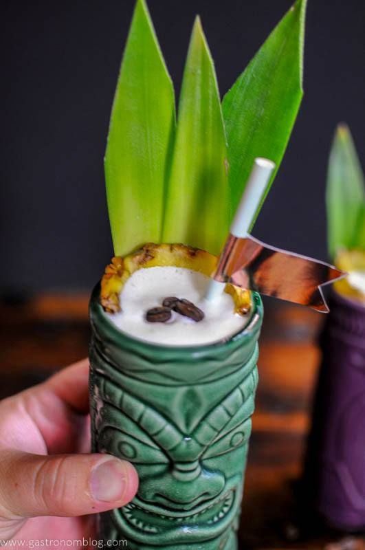 Hand holding green tiki mug with straw, coconut cream, coffee beans and pineapple fronds
