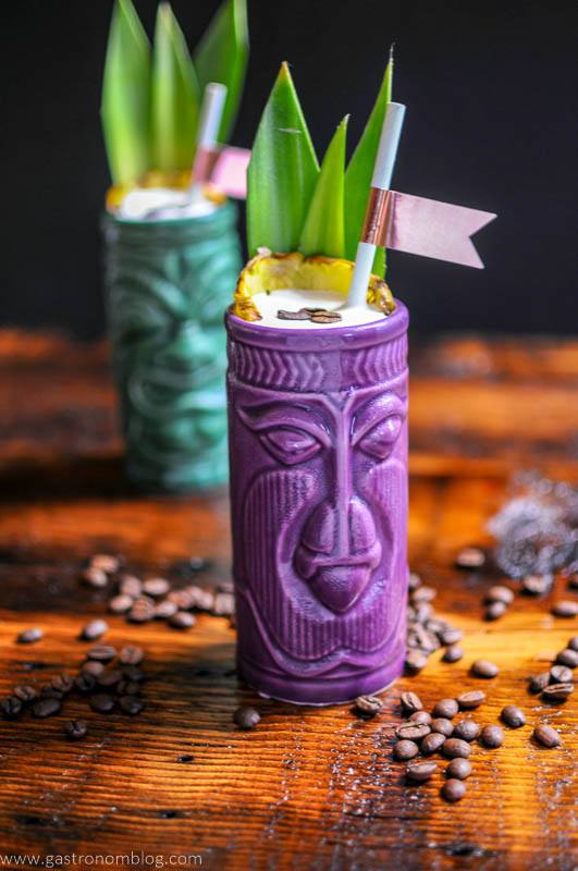 Purple and green tiki mugs with pineapple fronds, straw with gold flags. Coffee beans on wooden table