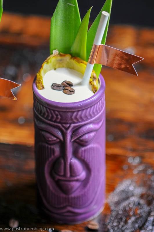 Purple tiki mug with coconut cream, coffee beans, pineapple fronds and straw with gold flag