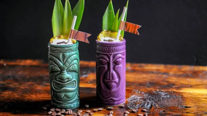 Green and purple tiki mugs with pineapple fronds and straws. Coffee beans on wooden table