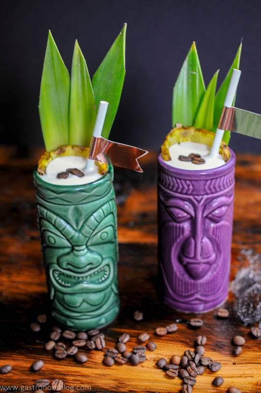 Green and purple tiki mugs with straws, pineapple fronds and coconut cream on top. Coffee beans on wooden table