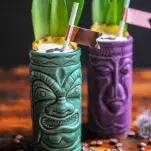 Green and purple tiki mugs with coconut cream and coffee beans on top, pineapple fronds and straws. Coffee beans on wooden table