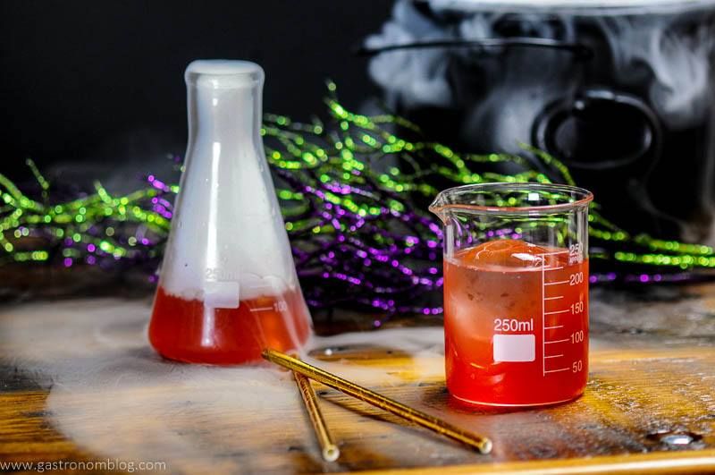 Red Halloween Cocktail in laboratory beakers. Gold straws and silver cocktail shaker. Cauldron in back with dry ice. 