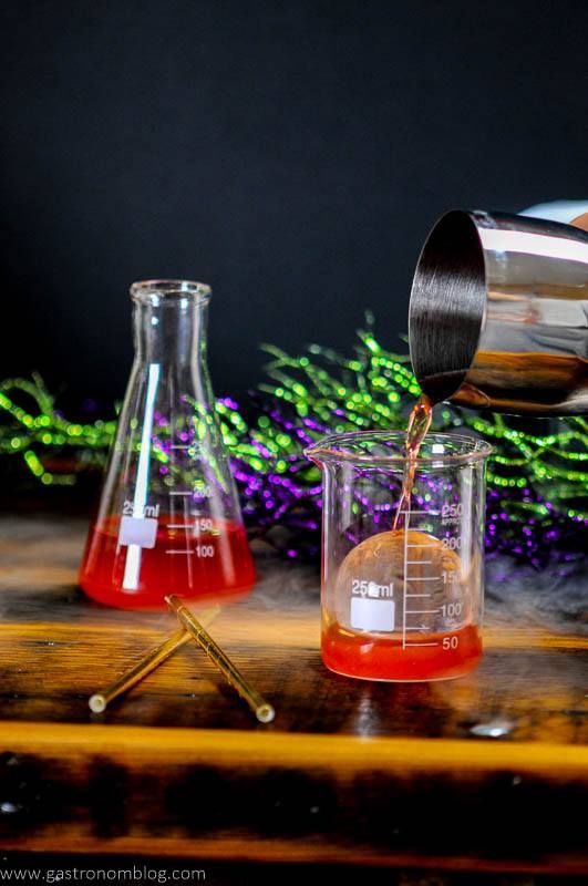 Red Halloween Cocktail being poured into laboratory beakers. Gold straws and silver cocktail shaker. 