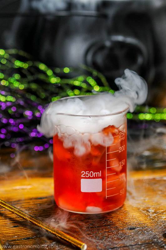 Red cocktail in laboratory beaker with dry ice smoke. Green and purple glitter sticks behind