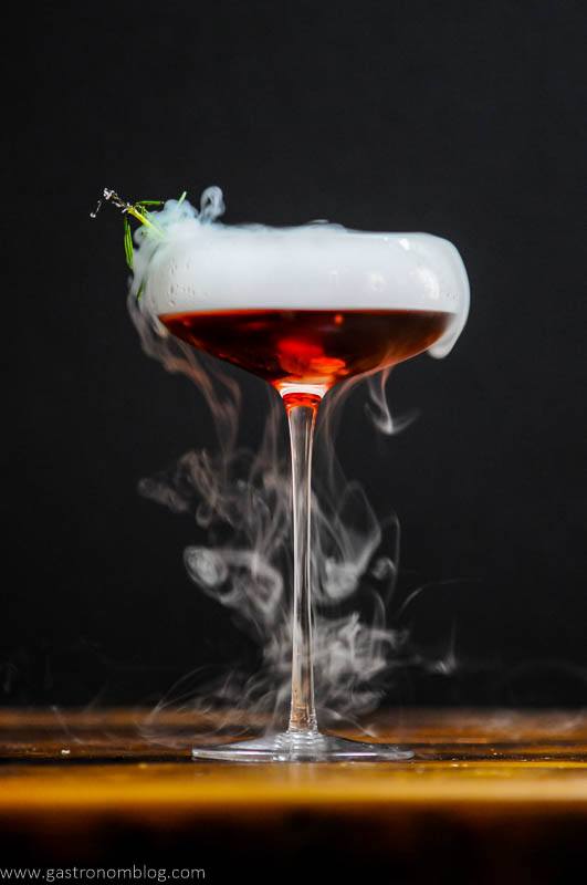 Dry ice boils out rolling fog from a red Cynar Negroni in a cocktail coupe