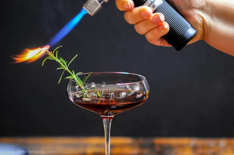 A culinary torch uses flame to set a rosemary sprig on fire in a cocktail coupe