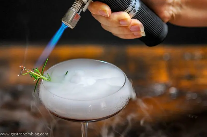 A culinary torch uses flame to set a rosemary sprig on fire in a cocktail coupe