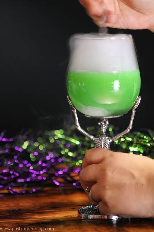 The Grindelwald Goblet - A green Halloween cocktail in a skeleton wine glass with dry ice smoke topping the drink while a hand moves it.