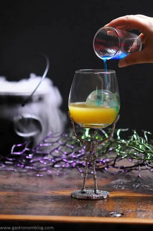 A wine glass supported by a metal skeleton holds a halloween cocktail while blue curacao is poured into it.