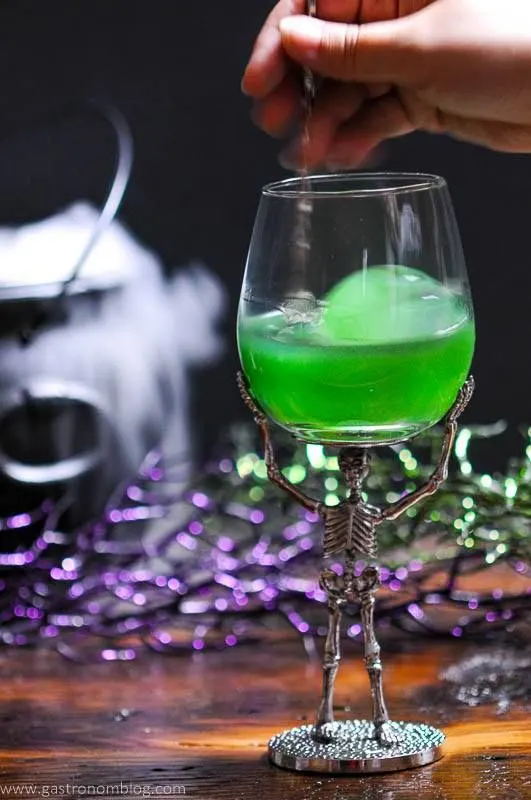 A green Halloween cocktail called the Grindelwald's Goblet is presented with a large circular ice cube in a skeleton based wine glass.