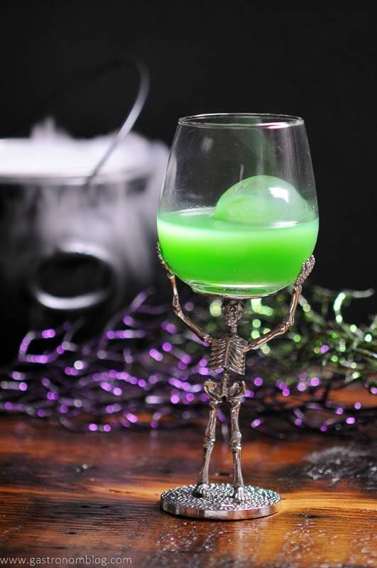 A green Halloween Cocktail called the Grindelwald's Goblet is presented with a large circular ice cube in a skeleton based wine glass.