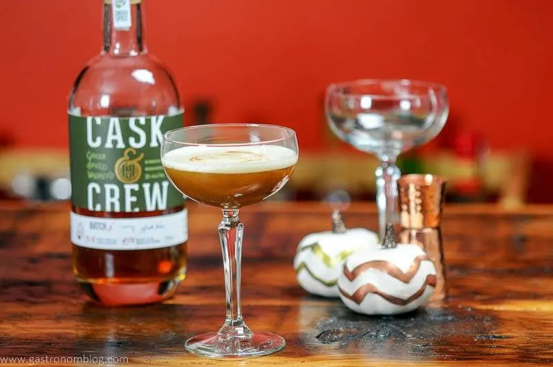 A closeup shot of a coupe glass filled with the Holiday Ginger Flip Cocktail on a wooden bar top along side a Cask And Crew Ginger Whiskey Botte.