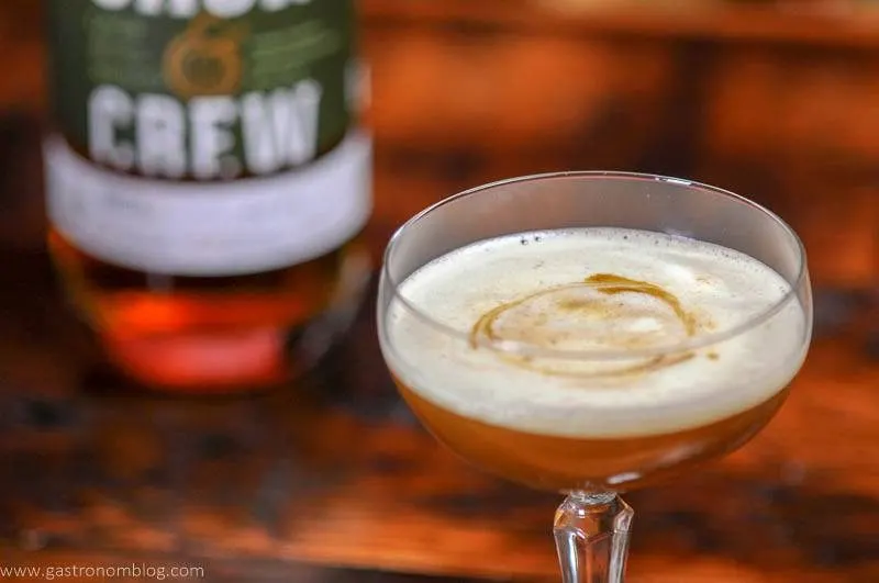 A coupe glass filled with a Holiday Ginger Fip whiskey cocktail is topped with a thick creamy foam and garnished with drops of bitters.