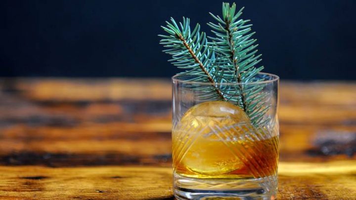 A rocks glass filled with a Pine Old Fashioned cocktail and a clear ice ball on a bar top.