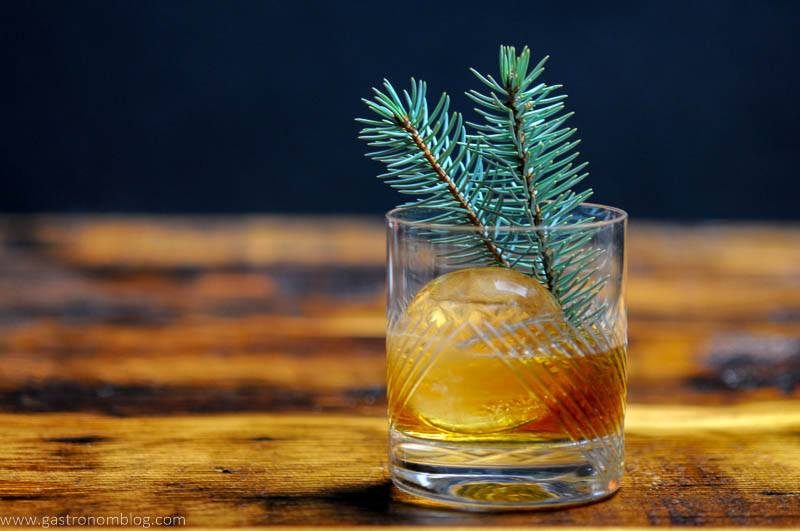 A rocks glass filled with a Pine Old Fashioned cocktail and a clear ice ball on a bar top.