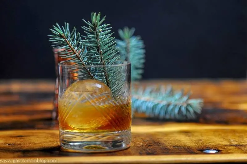 A hand cut crystal rocks glass filled with a Pine Old Fashioned with a clear sphere of ice and a pine branch garnish on a bar top.