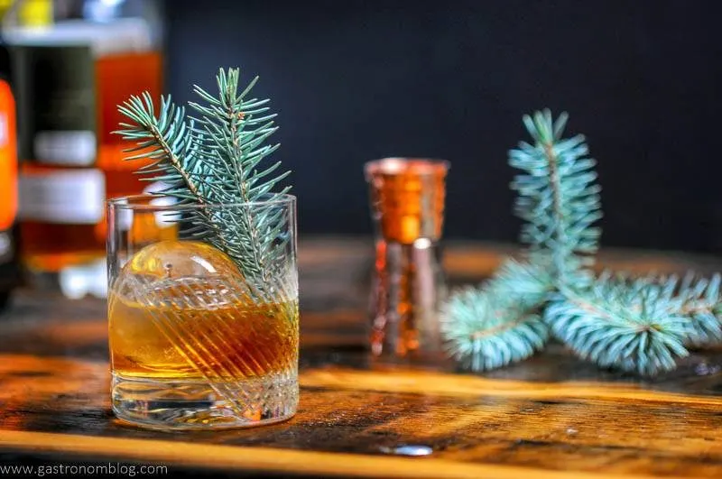 A rocks glass filled with a Pine Old Fashioned and a clear sphere of ice on a bar top and garnsished with a pine branch sits in front of a bunch of spirits bottles.