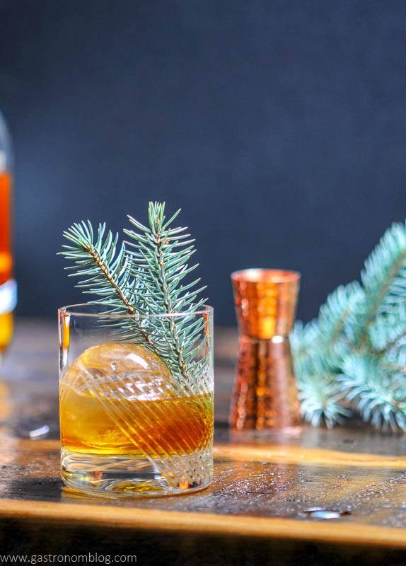 A rocks glass filled with a Pine Old Fashioned and a clear sphere of ice on a bar top.