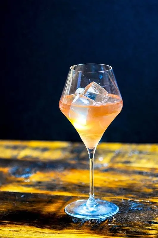 A tall wine glass sits on a wooden bar top filled with the Honey Bourbon and Pear Cider Cocktail, a pale orange colored cocktail featuring Barenjager Honey and Bourbon served on the rocks.