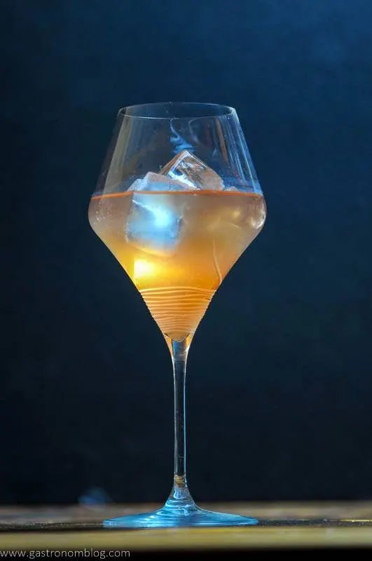 A tall wine glass sits on a wooden bar top filled with the Honey Bourbon and Pear Cider Cocktail, a pale orange colored cocktail featuring Barenjager Honey and Bourbon served on the rocks.