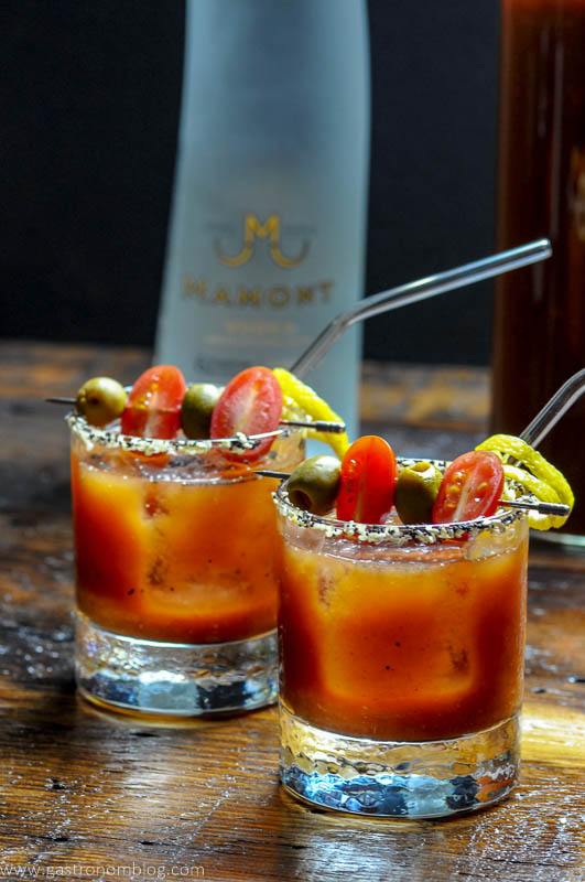 A pair of rocks glasses and a glass pitcher filled with a Bloody Mary cocktails sits on a wooden bar top with a bottle of Mamont Vodka alongside to celebrate National Bloody Mary Day.