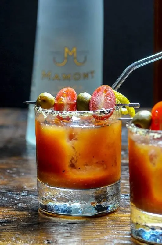 A pair of rocks glasses and a glass pitcher filled with a Bloody Mary cocktails sits on a wooden bar top with a bottle of Mamont Vodka alongside to celebrate National Bloody Mary Day.