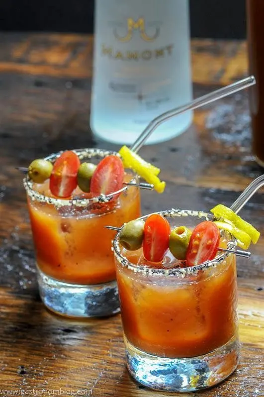 A pair of rocks glasses and a glass pitcher filled with a Bloody Mary cocktails sits on a wooden bar top with a bottle of Mamont Vodka alongside to celebrate National Bloody Mary Day.