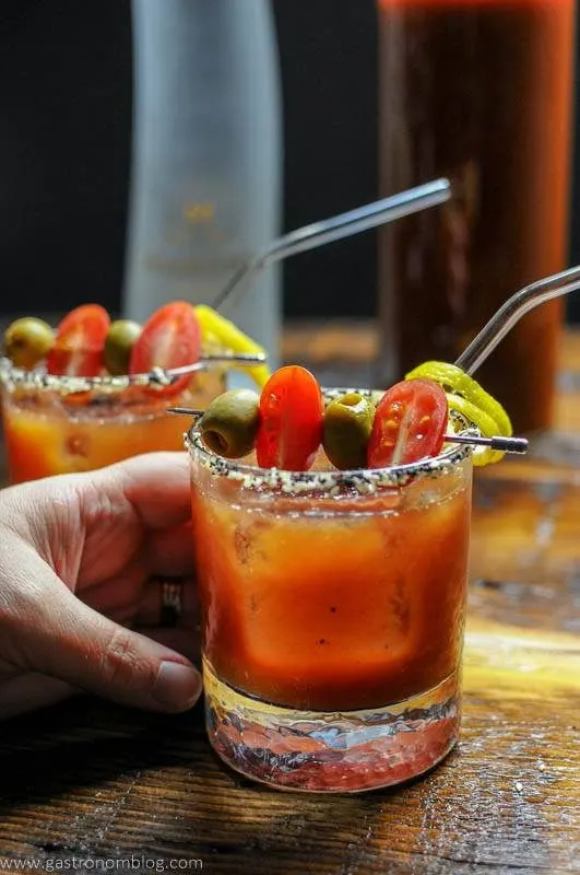 A hand grasps a rocks glass filled with a Bloody Mary Cocktail topped with olives and cherry tomatoes as a garnish.