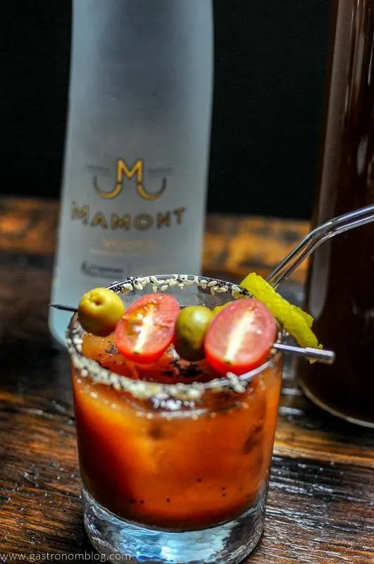 A rocks glass filled with a Bloody Mary cocktail sits on a wooden bar top with a bottle of Mamont Vodka alongside to celebrate National Bloody Mary Day.