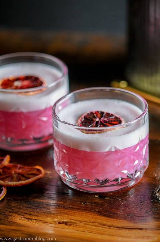 A rocks glass filled with a blood orange gin sour topped with white foam and a dried blood orange slice sits on a wooden bar top.