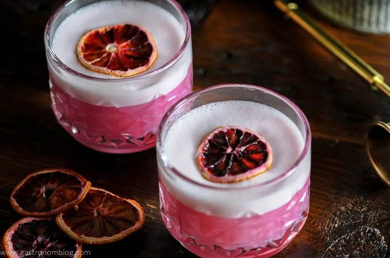 Two rocks glasses filled with a blood orange gin sour topped with white foam and a dried blood orange slice sits on a wooden bar top.