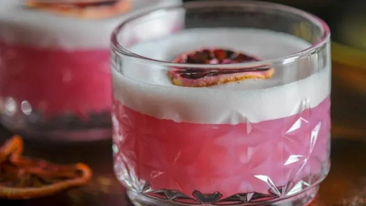 A rocks glass filled with a blood orange gin sour topped with white foam and a dried blood orange slice sits on a wooden bar top.
