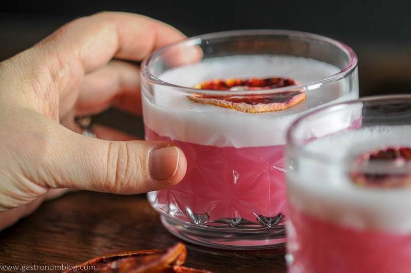 A hand grasps one of two rocks glasses filled with a blood orange gin sour cocktail.