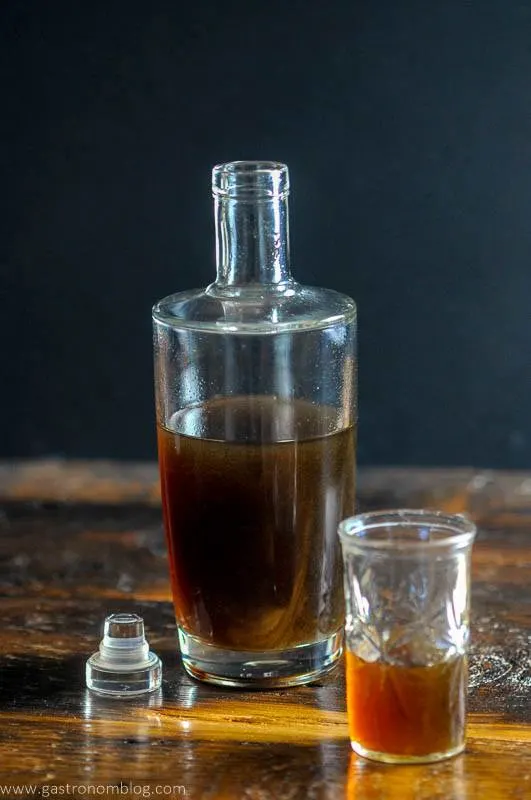 A bottle full of homemade banana liqueur sits on a wooden bar top next to a small shot glass with banana liqueur in it.