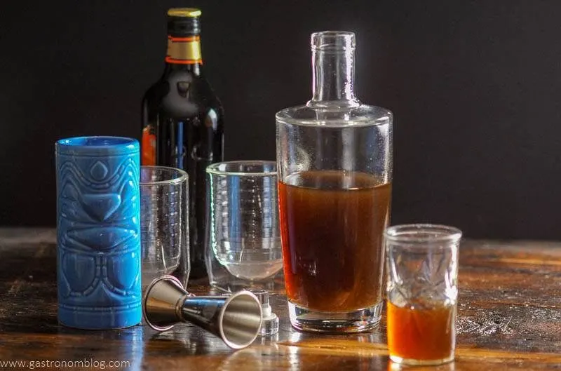 A bottle full of homemade banana liqueur sits on a wooden bar top next to a small shot glass with banana liqueur in it while tiki mugs sit alongside.