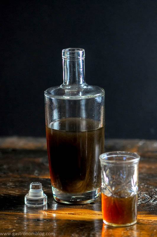 A bottle full of homemade banana liqueur sits on a wooden bar top next to a small shot glass with banana liqueur in it.